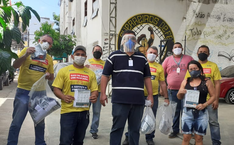 secretário e pessoas ao lado, todos de máscara, em rua segurando pacotes com máscaras e posando para a foto